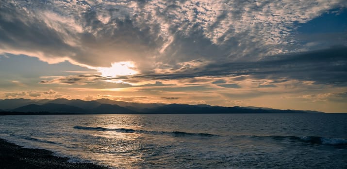 Waves gently lap the shore as the sun sets behind mountains, casting warm hues across the expansive sky.