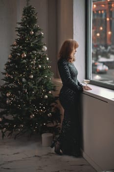 A beautiful elegant woman in a long dress next to the Christmas tree. Christmas and New Year holidays.