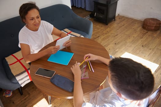 A mother and son interacting during a homeschooling session at a wooden table, showcasing learning and family bonding. The setting reflects a cozy and supportive educational environment.