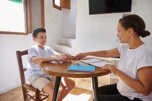 A mother and son are seated at a table, engaging in homeschooling activities with notebooks and pencils, sharing a joyful and educational moment together in a bright room.