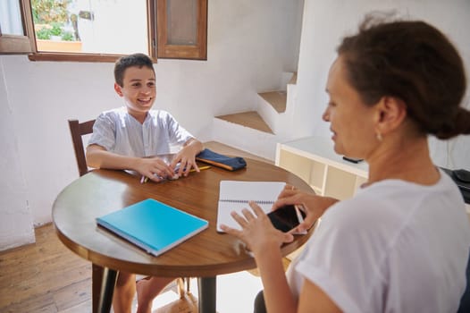 A mother and son engage in a joyful homeschool session at a wooden table, surrounded by notebooks and pencils. The bright room and open window convey a sense of warmth and focus.