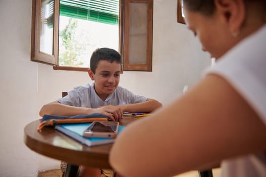 A young boy sits at a table with his mother, engaging in conversation in a cozy daytime setting. The room is warmly lit by sunlight streaming through open windows, creating a relaxed atmosphere.