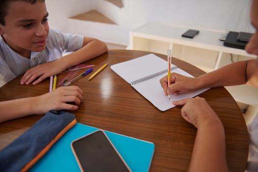 Two children are sitting at a table engaged in a study session. One is writing in a notebook while the other watches attentively. Pencils and school supplies are scattered on the table.