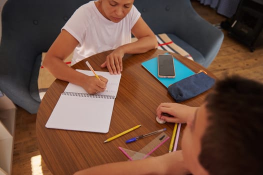 A mother helps her child with homework at a kitchen table, surrounded by school supplies. The scene captures family bonding, education, and study time in a cozy home setting.