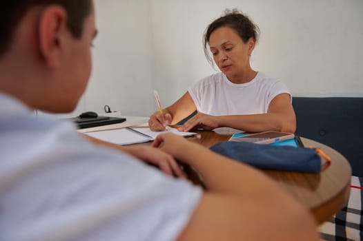 A woman in white shirt engages with a young boy during a home tutoring session. They sit at a round table, focused on learning. The scene conveys education, mentorship, and personal growth.