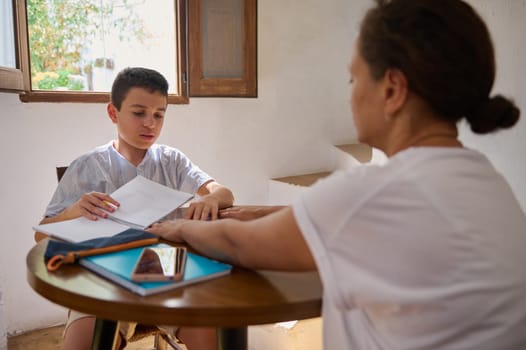 A mother guides her son through a homeschooling session at a wooden table, fostering learning in a sunlit room.