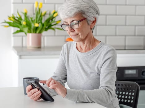 Grey-Haired Attractive Woman Searches For Information On Phone At Home At Table