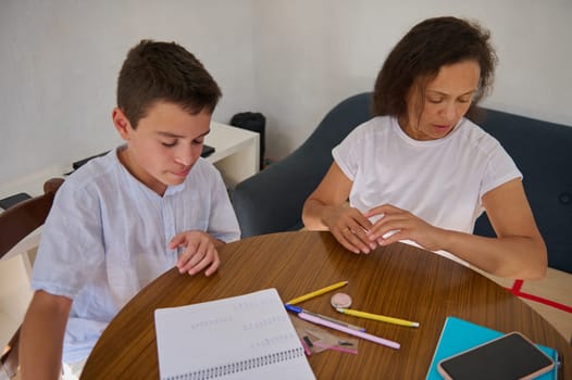 A mom guides her son through his homework at the kitchen table. Pencils, a notebook, and concentration are visible, fostering a learning environment.