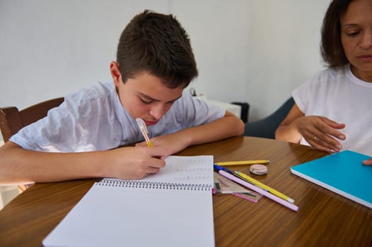 A young boy concentrates on his homework at a table, with school supplies scattered around, while a parent provides support and guidance nearby.