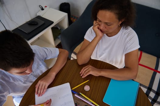 A mother sits at a table with her son, assisting him with math homework. The boy is concentrating on the worksheet, surrounded by pencils and notebooks, while she looks thoughtful and involved.