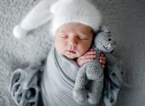 Newborn Baby Sleeps In Grey Wrap With Bunny Toy During Photoshoot