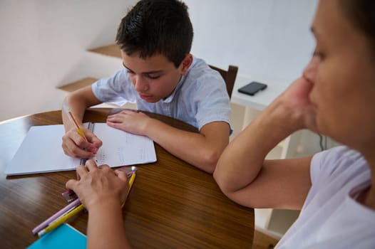 A caring mother assists her son with his homework at the dining table, providing guidance for his educational journey. The scene illustrates family support in learning.