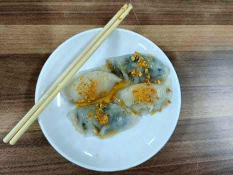 A plate of Choipan cakes from Indonesia sits on a wooden table. The cakes are white with green accents and are topped with a red sauce. Chopsticks are placed on the plate.