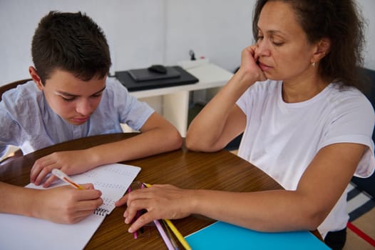 A mother helps her son with his homework at a wooden table. The boy is focused on solving math problems in a notebook while the mother supports him.