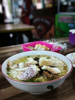 A bowl of Bubur Ikan Singkawang sits on a wooden table. The porridge is topped with chunks of fish and a red sauce. Chopsticks and other dishes are visible in the background.