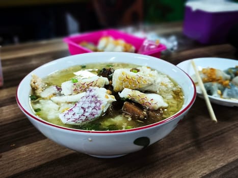 A bowl of Bubur Ikan Singkawang sits on a wooden table. The porridge is topped with chunks of fish and a red sauce. Chopsticks and other dishes are visible in the background.