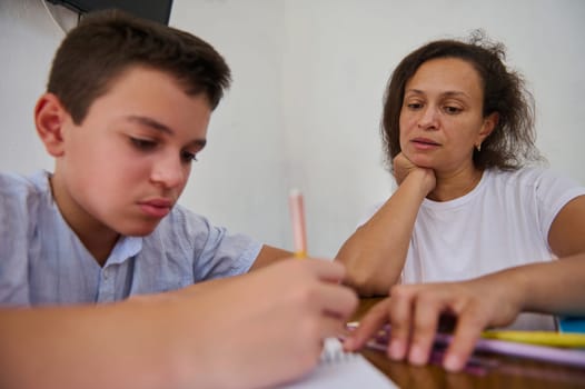 A mother attentively assists her son with his homework at the kitchen table. The boy focuses on writing while the mother guides him, displaying a scene of learning and support at home.