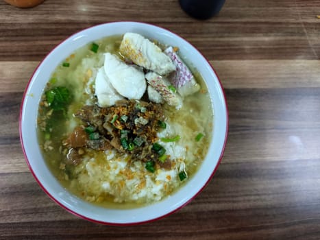 A bowl of Bubur Ikan Singkawang sits on a wooden table. The porridge is topped with chunks of fish and a red sauce. Chopsticks and other dishes are visible in the background.
