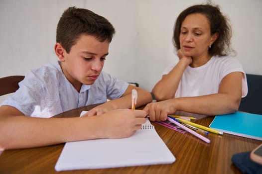 A focused boy works on homework under the guidance of his mother at the dining table, creating an engaging educational moment.
