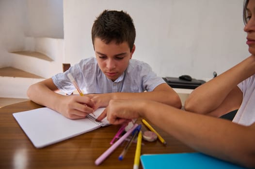 A young boy focuses intensely on his homework as an adult guides him, surrounded by colorful stationery on a wooden table.