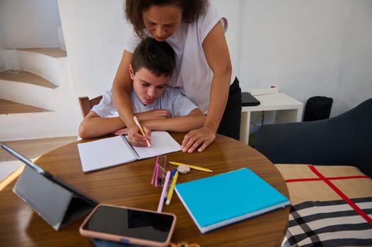 A mother lovingly assists her son with his homework at a wooden table, fostering learning and support. Tablets and notebooks are scattered, creating a cozy study environment.
