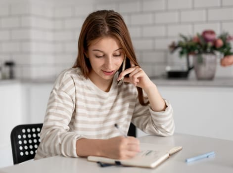 Young Girl Talks On Phone And Makes Notes In Notebook At Home