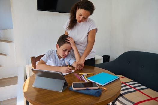 A mother assists her son with his homework while he writes at a table using a tablet. The scene conveys support, education, and family bonding in a cozy home environment.