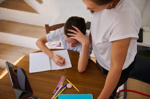 A parent assists their child with homework while sitting at a cozy wooden table. They are surrounded by stationery and a tablet, emphasizing a learning environment and supportive interaction.