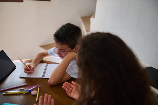 A young boy sits at a wooden table, focused on writing in a notebook, as a woman assists him with his homework. Various school supplies are scattered on the table.