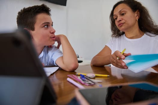 A mother helps her son with homework, offering guidance and support. The interaction promotes learning, education, and family bonding. Pens and notebooks are scattered on the table.