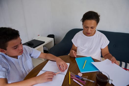 A mother and her son are seated at a wooden table, engaged in homeschooling. Notebooks, pens, and a cup of tea suggest a focused and educational atmosphere.