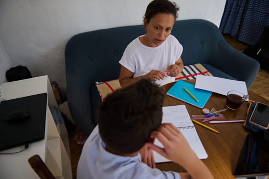 A mother assists her son with his homework, surrounded by notebooks and stationery on a wooden table. They engage attentively in their study session, emphasizing education and family support.