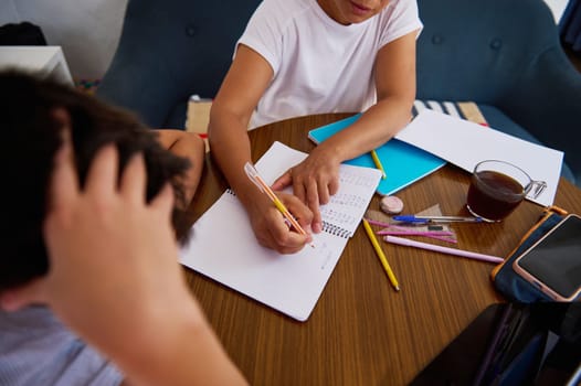 A mother helps her child with homework, showing patience and guidance. Pencils and notebooks are scattered on the table, illustrating a focused learning environment.