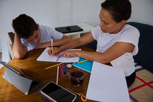 A mother patiently helps her child with homework at the dining table, surrounded by books and a tablet. The scene reflects concentration, support, and education.