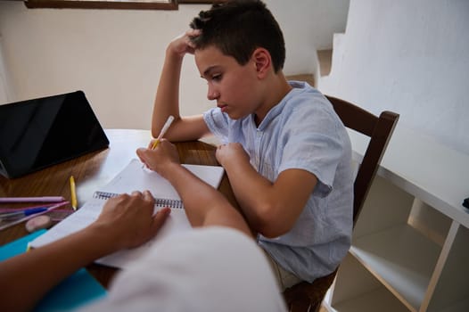 A young boy is deeply focused on his homework, writing in a notebook at a desk. The setting is calm, with a tablet and stationery nearby, emphasizing a studious atmosphere.