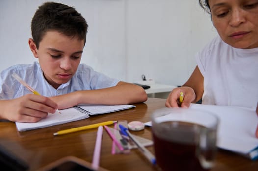 A mother and son sit at a table focusing on homework. The boy writes in a notebook while the mother provides guidance. Pencils and tea are scattered around, emphasizing a learning atmosphere.
