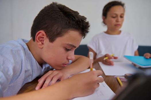 A young boy intensely focuses on his homework while sitting with a tutor. The image captures a learning environment emphasizing concentration, education, and guidance in a supportive atmosphere.