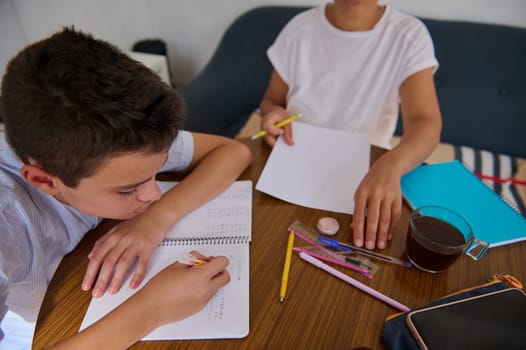 Two children are engaged in studying at a wooden table with notebooks, pens, and a cup of tea, creating a focused learning environment.