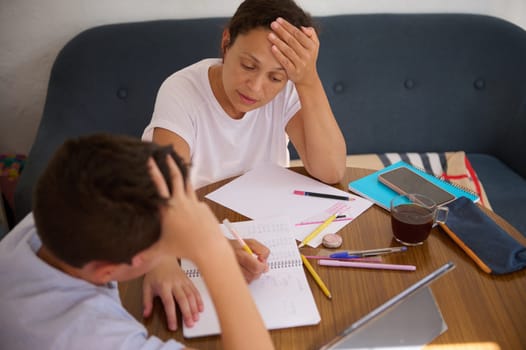 A mother and son sit at a table, focusing on homework together. The mother appears stressed while the son writes in a notebook. The scene conveys education, family support, and shared challenges.