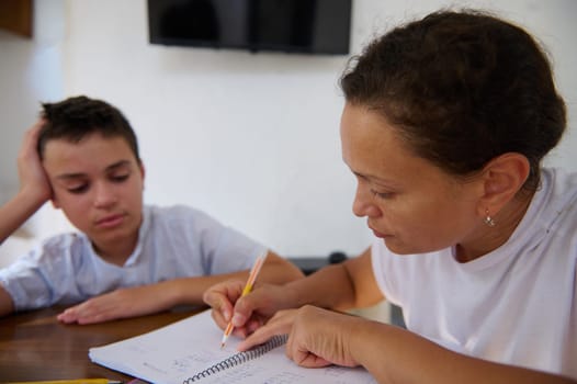 A mother guides her son through his homework during a study session at home. The boy appears slightly frustrated while the mother patiently explains concepts. The scene captures parental support.
