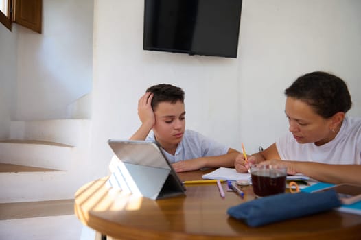 A mother assists her son with his homework at the dining table, focusing and guiding him. A tablet and stationery are scattered around, suggesting a productive learning environment.