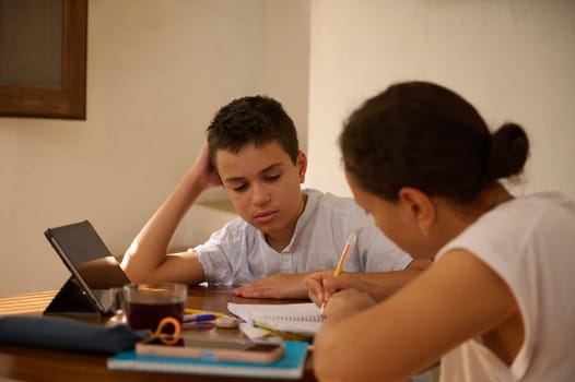 A mother and her son are focused on studying together at a table. The son is using a tablet while the mom assists with his homework, creating a supportive learning environment.