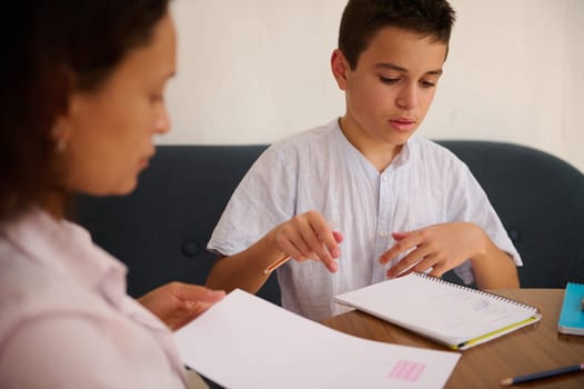 A mother guides her son through a focused homeschool lesson, emphasizing education and dedication. They are engaged with open books, fostering a supportive learning environment.