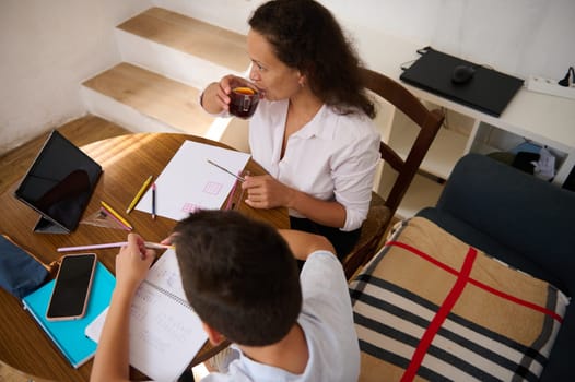 A mother helps her child with homework in a warm home setting, promoting learning and family bonding. They sit at a table with notebooks and a tablet, enjoying tea and study time.