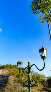 Elegant green street lamps with intricate designs stand in front of a vivid blue sky and lush hill. The scene exudes a blend of nature and historical architecture.