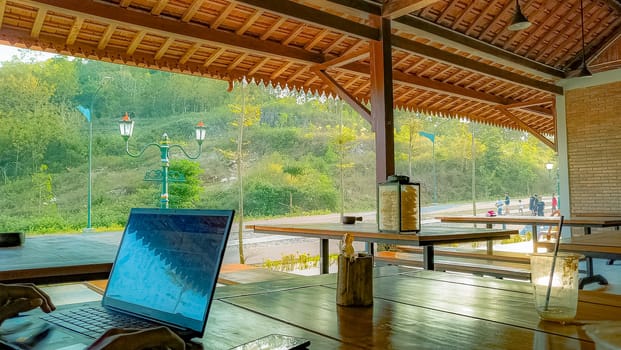 A serene outdoor workspace with a laptop on a wooden table, offering a peaceful view of nature and distant people enjoying the scenery.