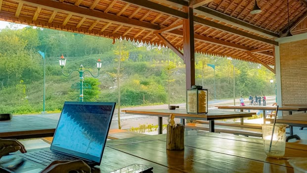 A serene outdoor workspace with a laptop on a wooden table, offering a peaceful view of nature and distant people enjoying the scenery.