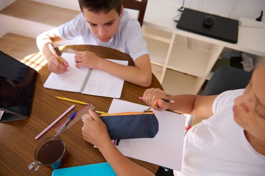 A parent and child study together at a wooden table, surrounded by notebooks and pencils. The tablet and coffee mug create a cozy homeschooling atmosphere, encouraging focus and learning.