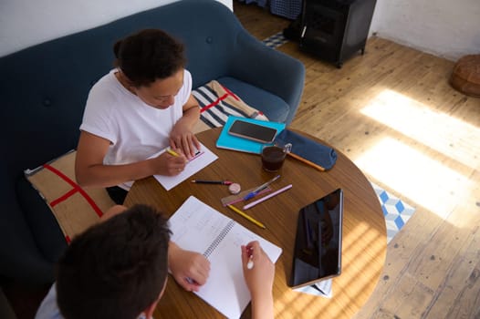 Two individuals work together at a cozy home workspace, surrounded by notebooks, tablets, and stationery. Sunlight casts patterns on the wooden floor, creating a warm and focused atmosphere.