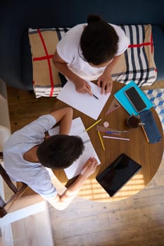 Two students working together at home on a project. Overhead view of a table with papers, tablets, and stationery, showing focused teamwork and learning.